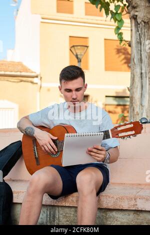 Creative male musician sitting on stone bench on street and reading notes while preparing for playing guitar Stock Photo