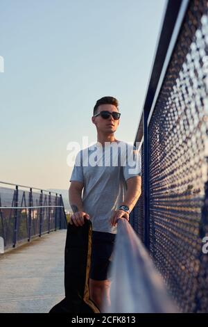 Confident male musician standing with guitar in case on bridge near railing and looking away Stock Photo