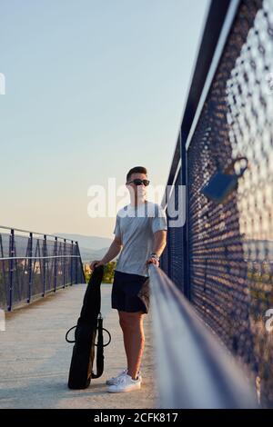 Confident male musician standing with guitar in case on bridge near railing and looking away Stock Photo