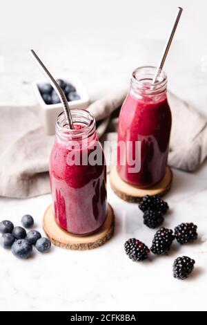 Berry smoothie and metal straws on glass bottles on wood slices over a white marble surface. High angle, vertical image. Stock Photo