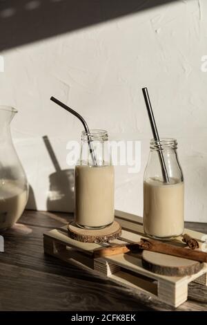Glass bottles filled with healthy homemade sweet milkshake with cinnamon placed on wooden table against wall with shadow of monstera leaves Stock Photo