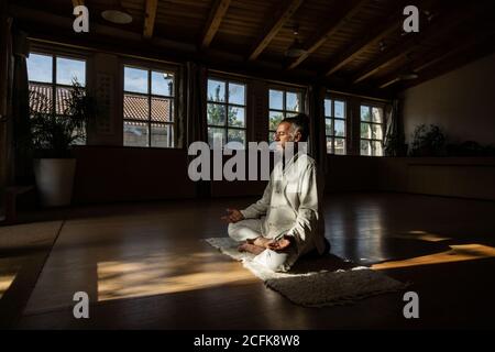 Full body side view of focused bearded ethnic senior male practicing chi kung meditation in lotus position in dark studio Stock Photo