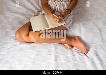 Blurred barefoot female sitting on bed and making notes in diary in cozy room on weekend day at home Stock Photo