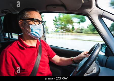 Middle aged senior man in medical mask driving modern car along road while looking forward Stock Photo