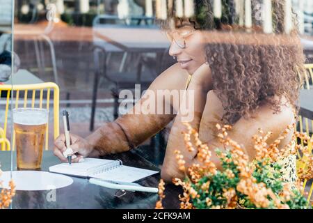 Side view of black self employed woman sitting at table in cafe and working remotely while writing in notepad