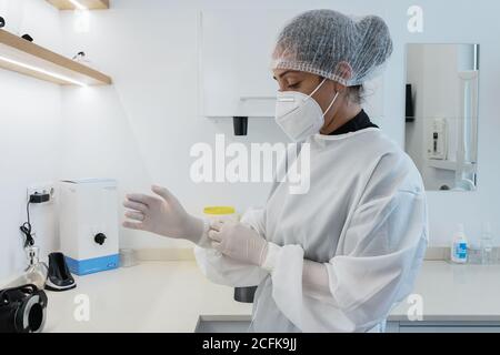 Side view of young female medical worker in white uniform and mask putting on latex gloves while preparing for treatment in modern clinic Stock Photo