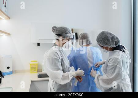 Female medical workers in protective masks and face shields helping each other to get dressed while preparing for medical procedure in hospital Stock Photo