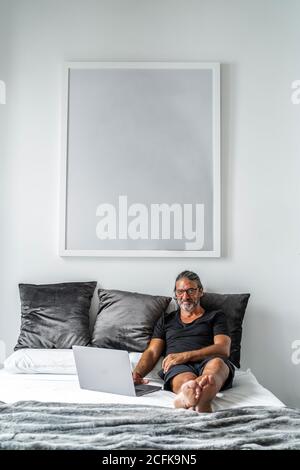 Smiling middle aged male entrepreneur sitting on bed and typing on laptop while working remotely on online project looking at camera Stock Photo