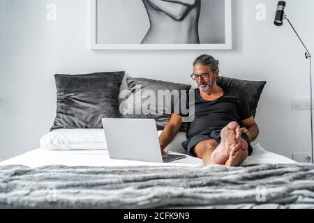 Smiling middle aged male entrepreneur sitting on bed and typing on laptop while working remotely on online project Stock Photo