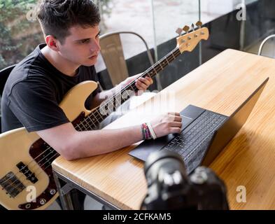 From above of focused young male sitting at table and watching video tutorial on laptop while learning to play music on bass guitar Stock Photo