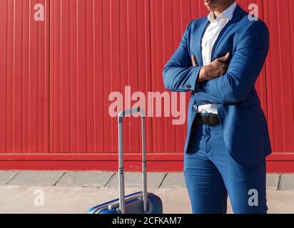 Crop unrecognizable black businessman in classy blue suit standing with arms crossed near suitcase against red wall Stock Photo