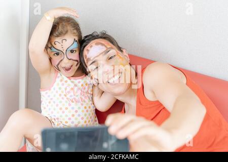 High angle of loving mother hugging cute little girl with painted face and taking photo on smartphone while having fun at home Stock Photo