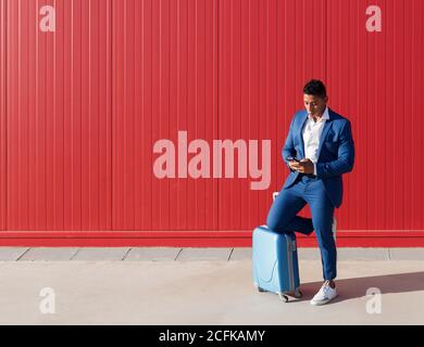 Full body of young African American man in elegant blue suit with luggage messaging on mobile phone while standing against red wall on street Stock Photo