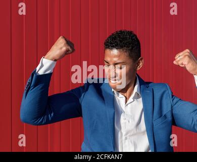 Confident satisfied young African American businessman in formal outfit celebrating success against red wall Stock Photo