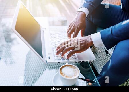 Side view of crop anonymous black businessman in formal suit sitting at table with cup of coffee and typing on laptop while working in cafe Stock Photo