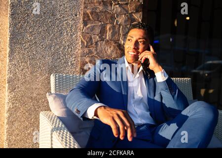 Young successful well dressed black businessman in elegant suit sitting in chair and having pleasant conversation on mobile phone Stock Photo