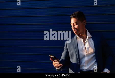 Happy young African American male manager in elegant formal clothes reading message on smartphone and smiling  while standing against dark blue wall on street Stock Photo