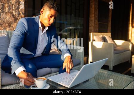 Young African American male entrepreneur in formal wear working on laptop and thinking about financial problem while sitting in lounge zone of modern cafeteria Stock Photo