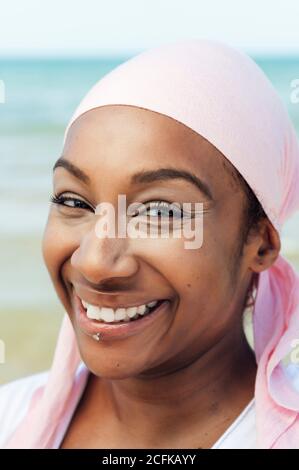 Determined ethnic female with headdress and piercing standing on beach on background of sea and looking at camera Stock Photo