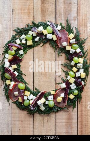Top view creative Christmas wreath made with green herbs and decorated with fresh grape and chopped cheese and ham arranged on wooden table Stock Photo