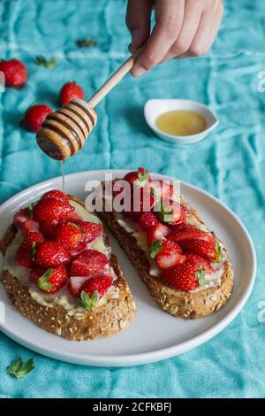 From above unrecognizable Woman pouring honey to yummy sweet bread toasts with fresh chopped strawberries and honey served on plate on blue tablecloth Stock Photo