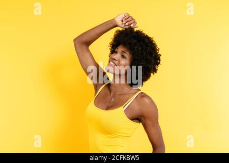 Positive African American female with Afro smiling and raising arm while standing against yellow background Stock Photo