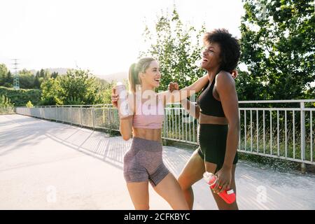 Happy young multiracial female friends in sportswear with bottles of water in hands chatting and laughing while having rest after fitness workout in summer park Stock Photo