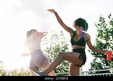 Low angle of happy young multiracial sportswomen in activewear with bottles of water having fun and jumping during fitness workout in park in summer day Stock Photo