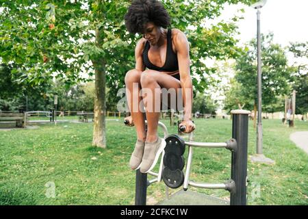 Low angle of positive young curly haired black sportsWoman doing push ups on parallel bars in summer park Stock Photo