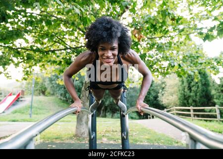 Low angle of positive young curly haired black sportsWoman doing push ups on parallel bars in summer park Stock Photo