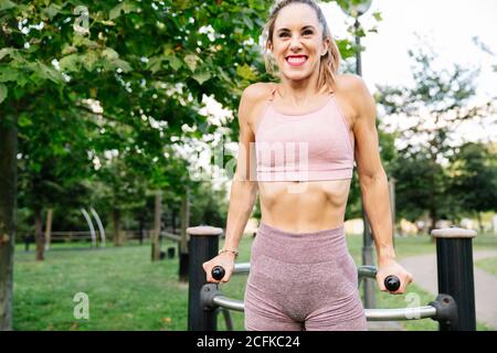 Positive young slim female in sportswear doing exercises on sports equipment during fitness workout in summer park Stock Photo