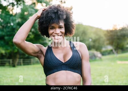 Young African American sportsWoman with curly hair wearing black top looking at camera with smile while standing in green park during workout Stock Photo