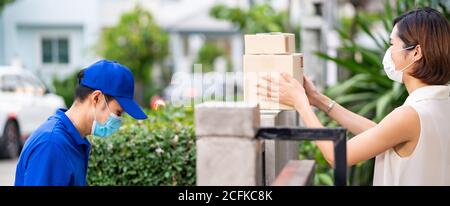 Panoramic side view asian woman with face mask customer take shopping packages from fence post that delivery man put them on for contactless delivery. Stock Photo