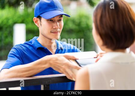 Asian deliver man handling package box to asian woman in front of house and female customer sign electronic signature to portable mobile device. Packa Stock Photo