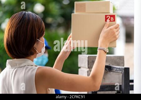 Asian woman with face mask customer take shopping packages from the fence post of customer house that delivery man put them on for contactless deliver Stock Photo