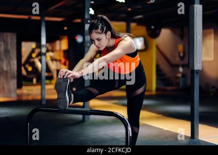 Full body young sporty female in fitness outfit performing stretching exercise near metal bar while warming up during training in gym Stock Photo