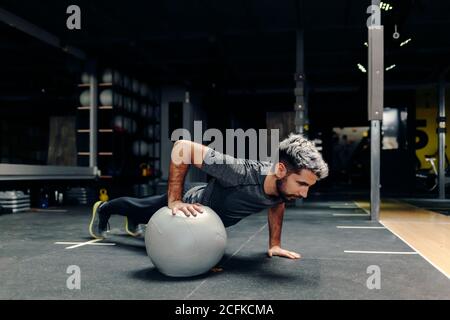 Full length of strong determined athletic male doing plank exercise using one hand with medicine ball during workout in gym Stock Photo