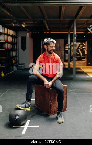 Full body positive young bearded male in sportswear sitting on wooden box near medicine ball and smiling while having break during training in modern sports club Stock Photo
