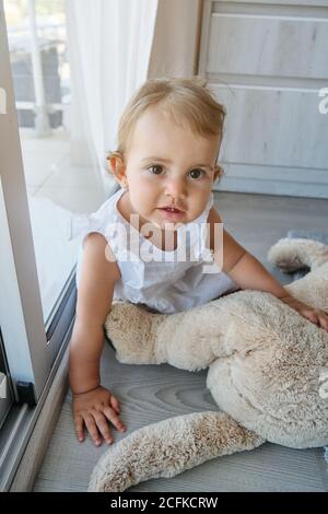 Portrait photo of a little girl in a white dress facing the camera with a teddy bear of a dog next to a windows of a house Stock Photo