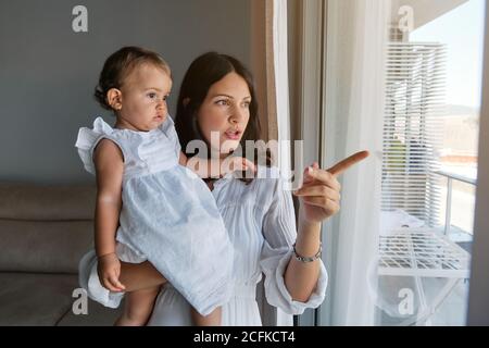 Mother with a little girl in white dress in her arms in front of a window pointing outwards in a house Stock Photo