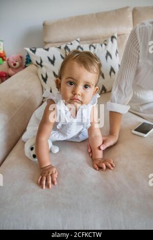 Vertical photo of a little girl in a white dress crawling on a sofa with her mother and a mobile phone next to her at home Stock Photo