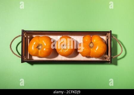 Ttop view three ripe yellow farm tomatoes lying in small rectangular wooden box in center of green background. Stock Photo