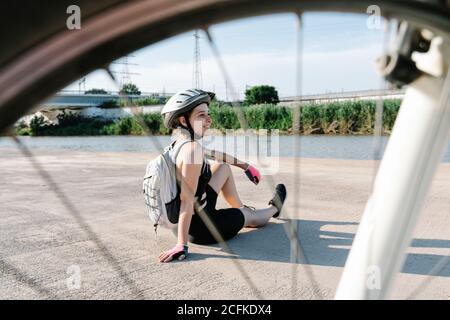 High angle side view of happy young Woman in sportswear and helmet with backpack sitting on paved ground and resting after riding bike in summer day Stock Photo