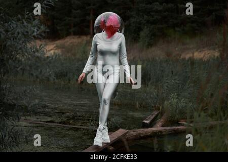 Happy young red haired female astronaut in silver suit and glass sphere helmet looking away while walking in field near forest in summer day Stock Photo