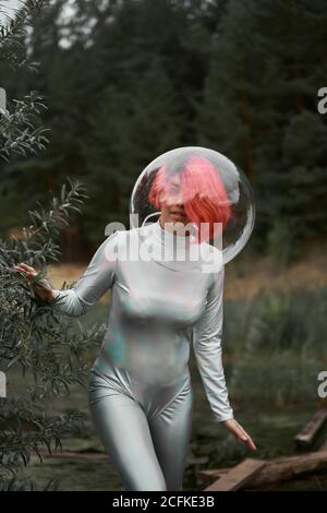 Happy young red haired female astronaut in silver suit and glass sphere helmet looking away while walking in field near forest in summer day Stock Photo