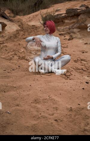 Positive young female with dyed red hair wearing silver space suit and glass helmet and looking away while sitting on rocky formation Stock Photo