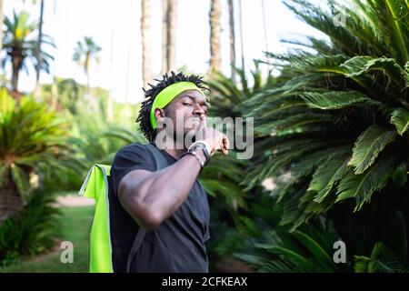 Young African American male athlete in sportswear and headband making shh gesture while standing against green tropical plants on street