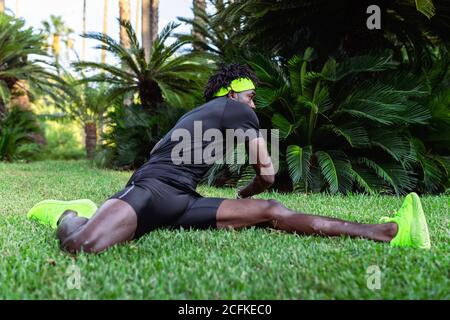 Young African American sportsman in stylish activewear sitting on green grass and doing stretching exercises after running in park