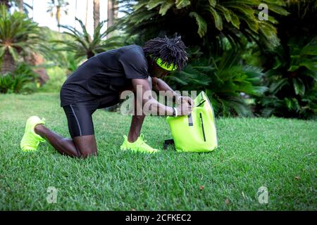 Side view of confident young African American male athlete with backpack in trendy sportswear getting ready to train in park with green tropical trees Stock Photo