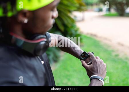 Stylish young African American male runner with headphones on neck checking sport results on fitness tracker during training in green tropical park Stock Photo